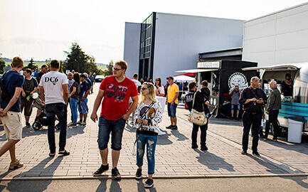 Menschenmenge bei sonnigem Wetter auf einem Event vor einem modernen Gebäude mit Foodtruck.