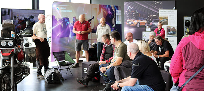 Gruppe von BMW Motorrad-Enthusiasten bei BMW Motorrad on Tour im Showroom mit ausgestellten Motorrädern und Infotafeln im Hintergrund.