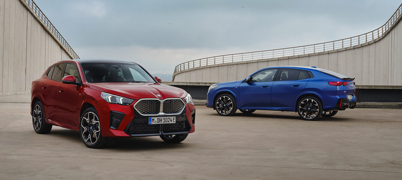 Zwei BMW iX2 Modelle in Rot und Blau auf einer modernen Betonfläche mit architektonischem Hintergrund.