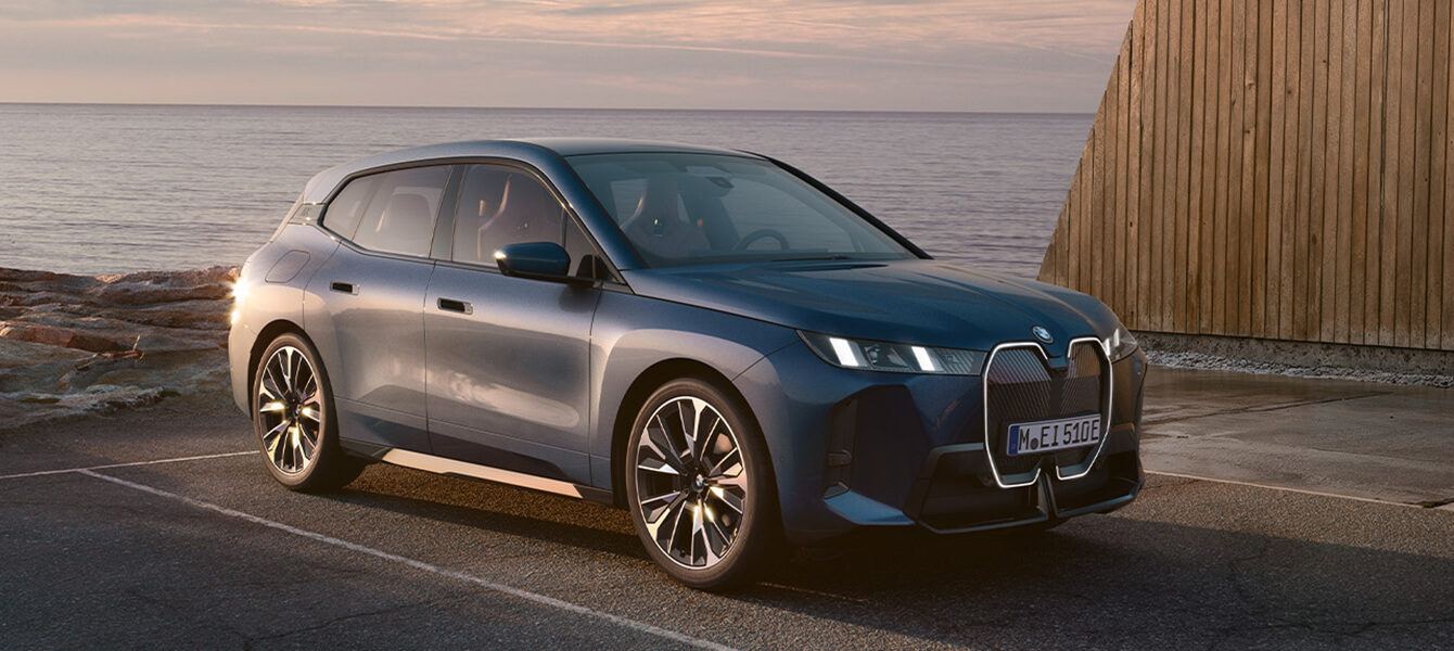BMW iX in blauer Lackierung vor moderner Architektur und malerischer Küstenlandschaft.