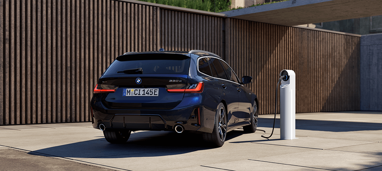 Heckansicht eines BMW 330e Touring Plug-in-Hybrid beim Laden an einer modernen Wallbox vor stilvoller Holzfassade.