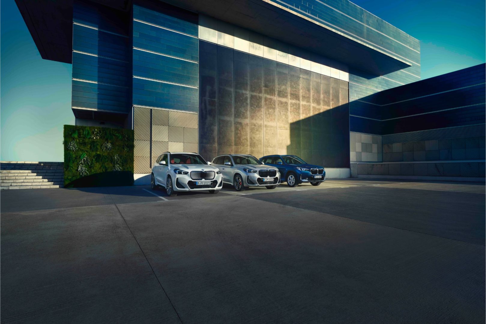 BMW X1, X1 als Hybrid und iX1 Modelle geparkt vor moderner Architektur. Erleben Sie den BMW X1 Modelle bei Ihrem Autohaus Kühnert.