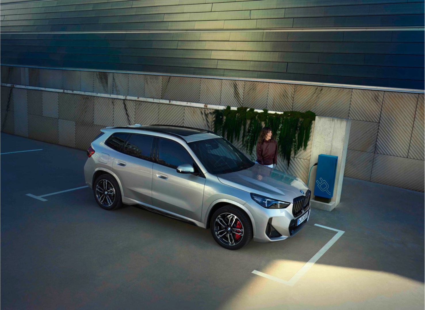 BMW iX1 beim Laden an Wallbox vor moderner Wandfassade. Erleben Sie den BMW iX1 bei Ihrem Autohaus Kühnert.