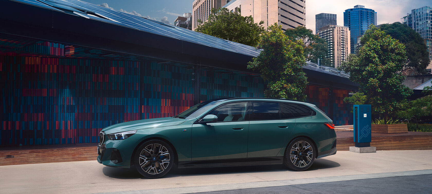 BMW 5er Touring Plug-in-Hybrid in grün, seitlich vor moderner urbaner Kulisse mit Ladeinfrastruktur.