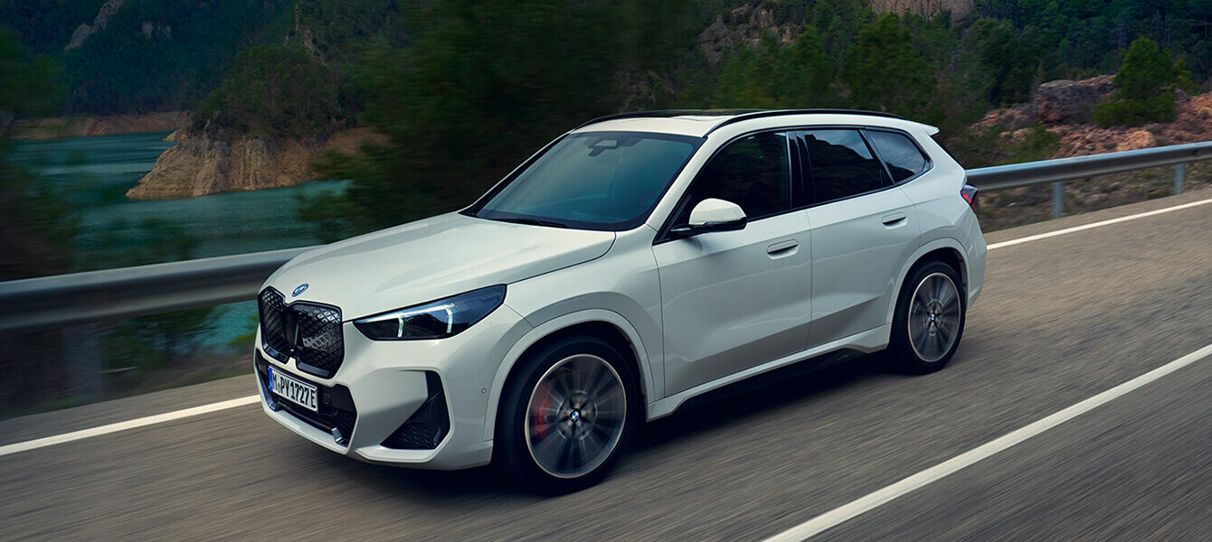 Weißer BMW iX1 fährt dynamisch auf einer kurvigen Landstraße entlang eines Sees mit bewaldeten Bergen im Hintergrund.