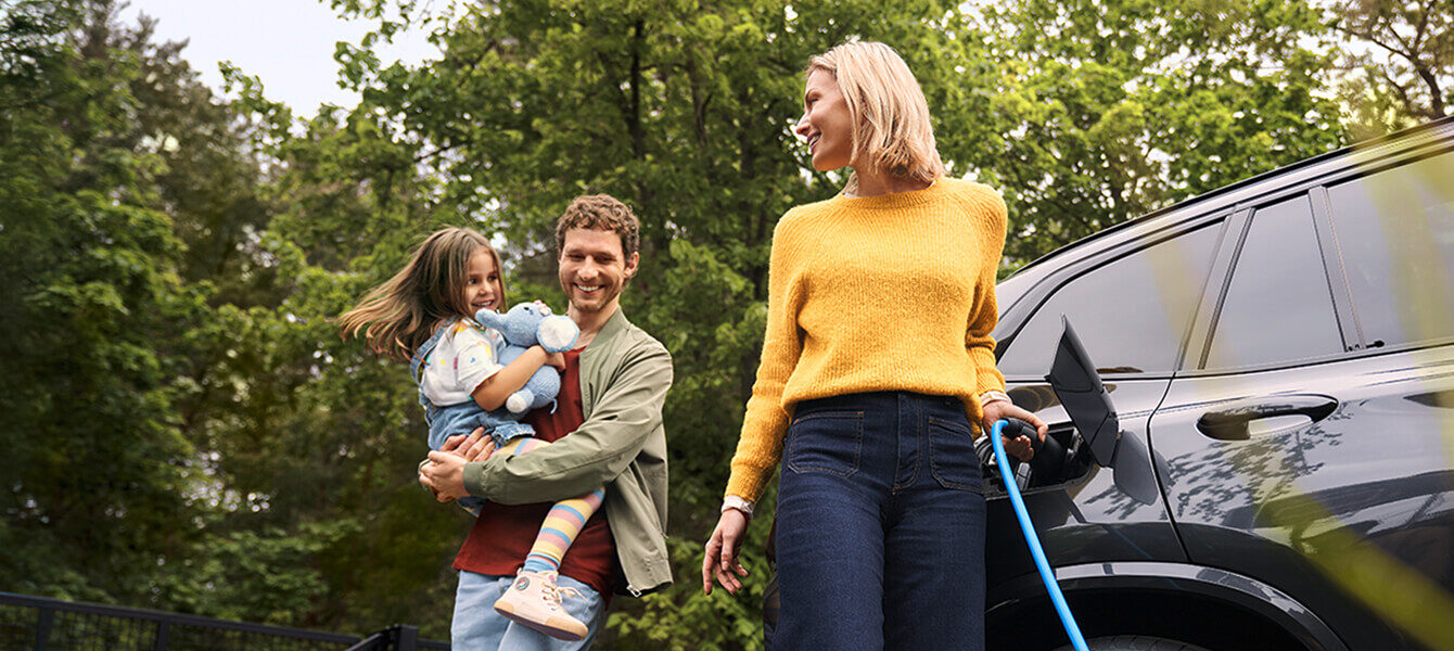 Familie lädt gemeinsam ein Elektroauto im Grünen, Eltern und Kind stehen neben dem Fahrzeug.