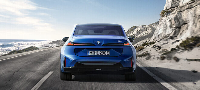 Blauer BMW i3 Neue Klasse von hinten auf einer Küstenstraße mit schmalen LED-Rückleuchten und sportlichem Heckdesign.
