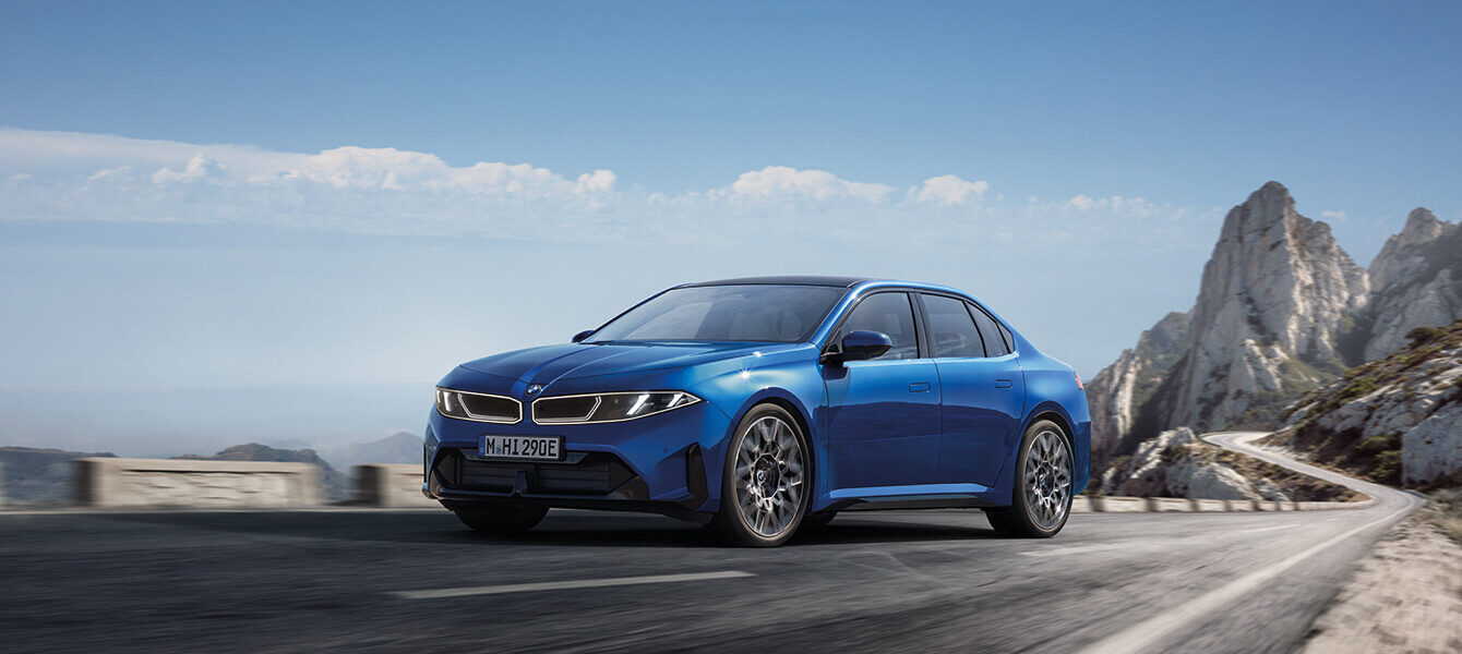 Blauer BMW i3 Neue Klasse in dynamischer Dreiviertelansicht auf einer Bergstraße mit felsiger Landschaft.