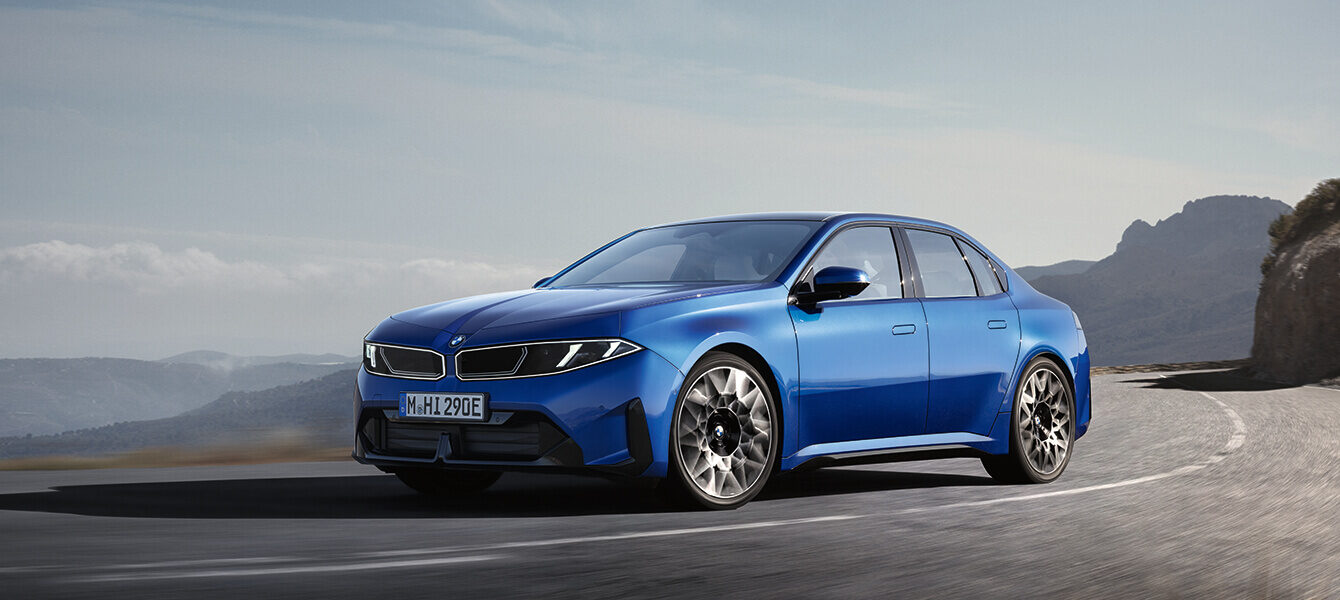 Neuer vollelektrischer BMW i3 Neue Klasse in Blau auf einer kurvigen Bergstraße, aufgenommen aus schräger Frontperspektive.